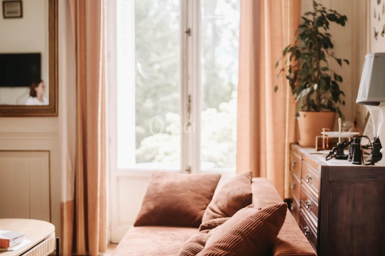 Canapé en velours brun devant une grande fenêtre lumineuse, entouré de rideaux beiges et de meubles en bois.