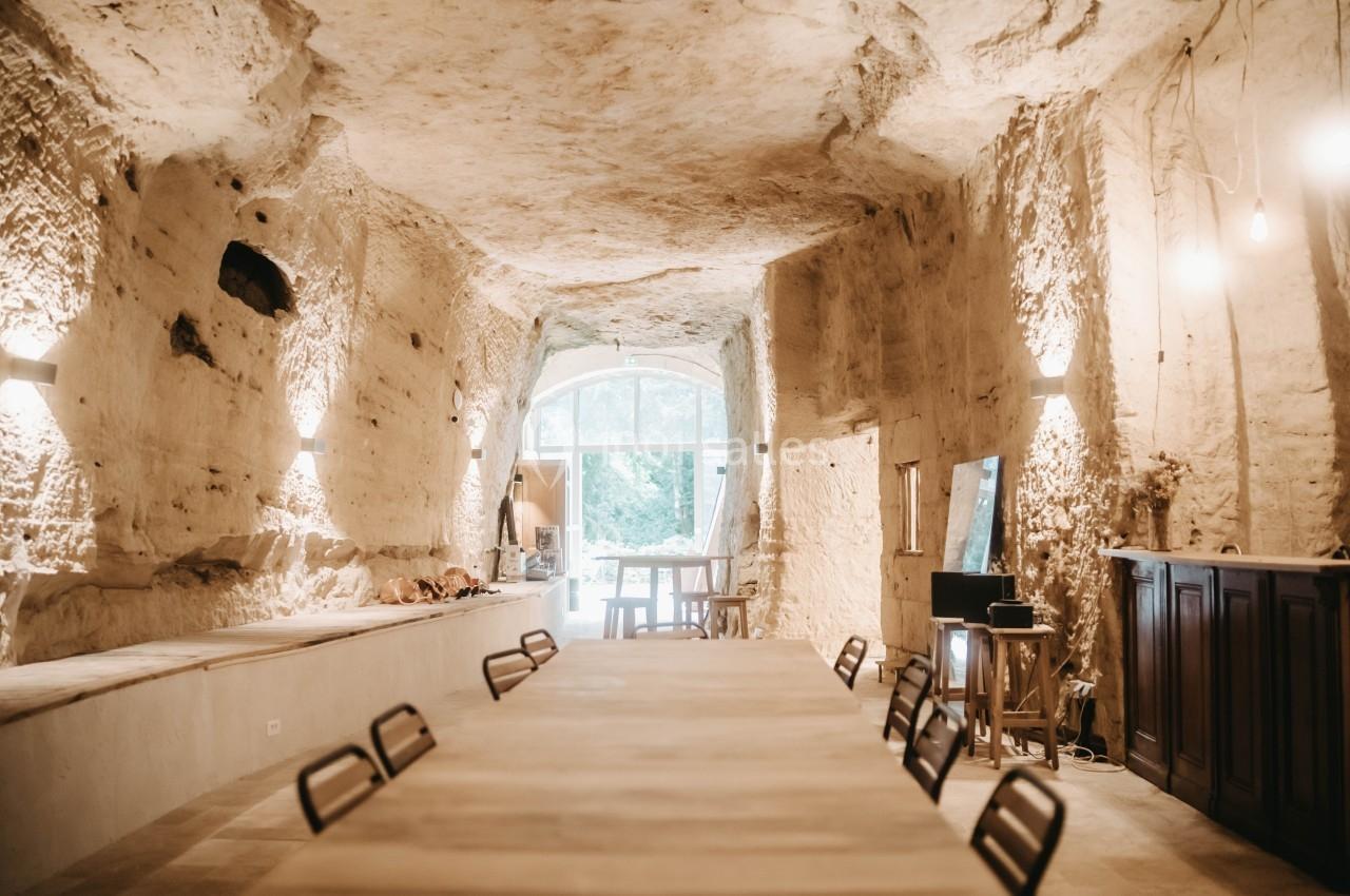 Salle voûtée en pierre naturelle avec une grande table en bois, éclairée par des lampes murales et une lumière naturelle.