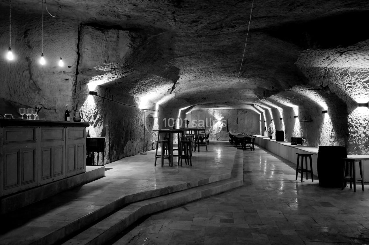 Salle voûtée en pierre avec éclairage tamisé, bar en bois à gauche et tables hautes dispersées.