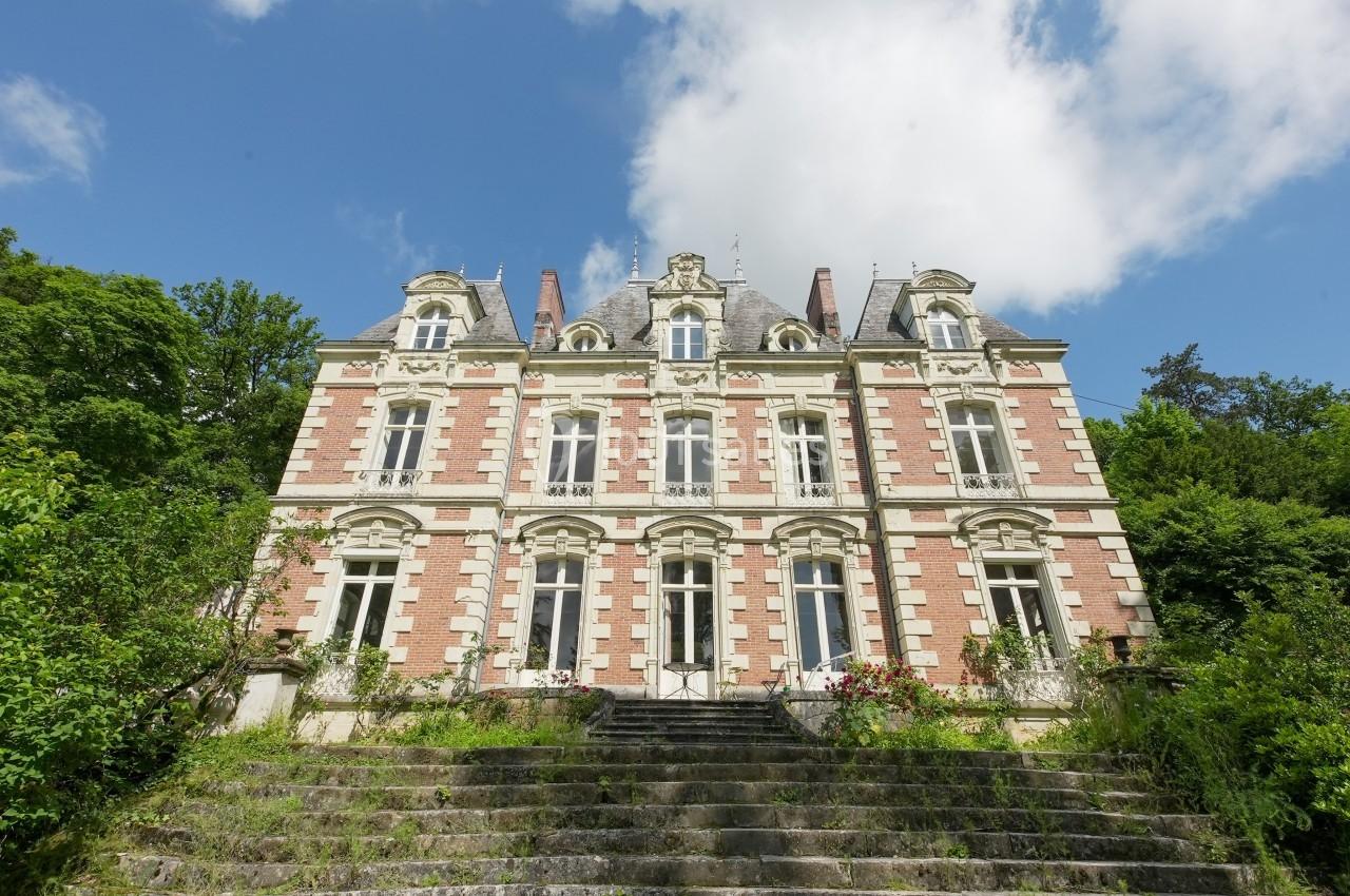 Façade d'un manoir en briques rouges et blanches, entouré de verdure, sous un ciel partiellement nuageux.