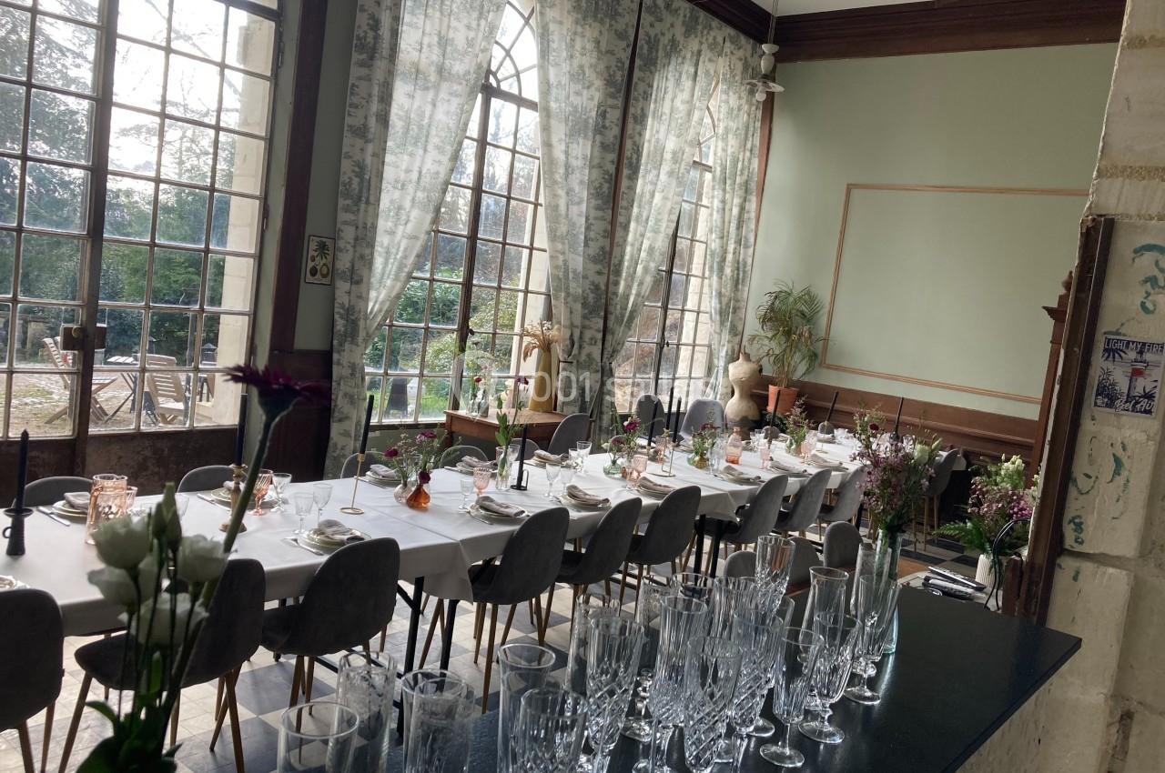 Salle lumineuse avec de grandes fenêtres, table dressée pour un repas, décorée de fleurs et entourée de chaises grises.