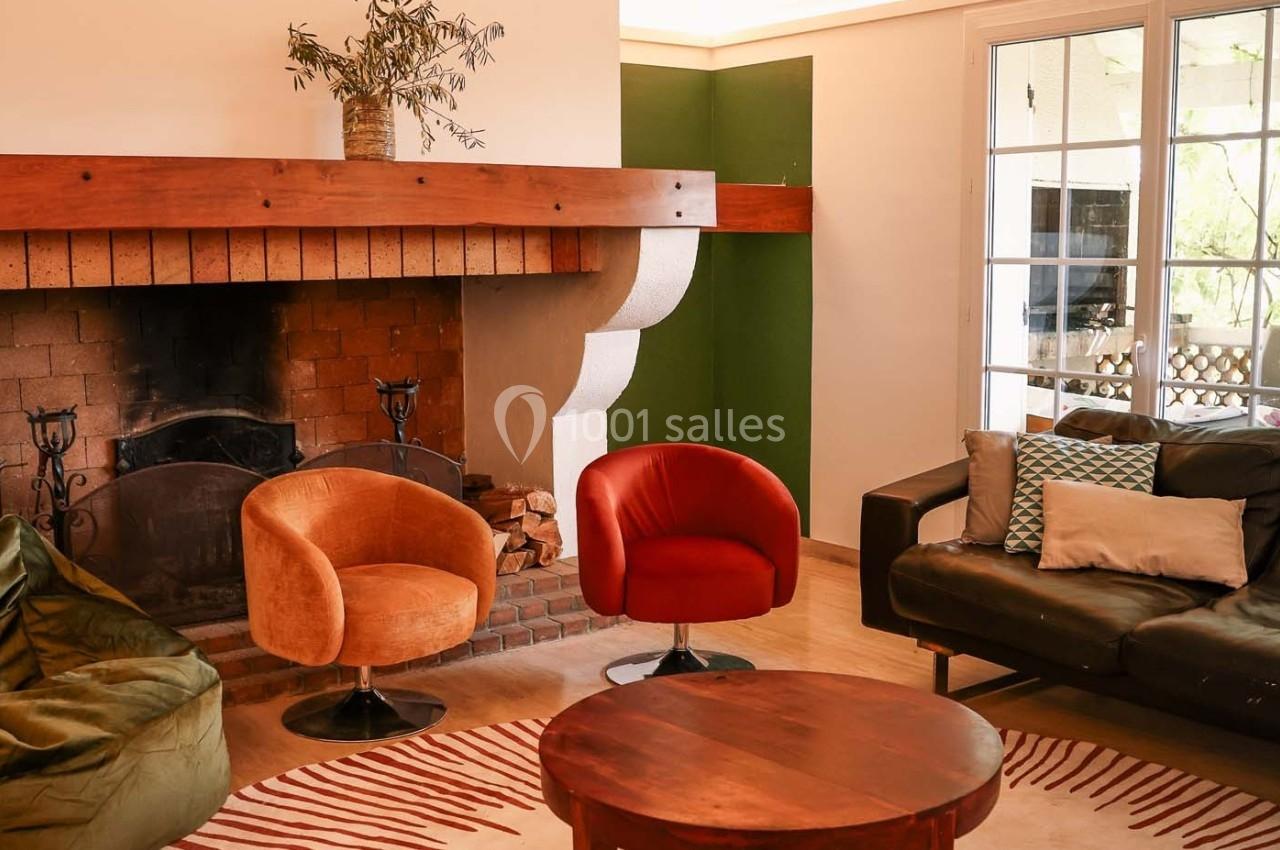 Salon avec cheminée en briques, fauteuils colorés, canapé en cuir et table basse ronde sur un tapis rayé.