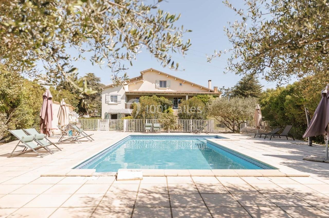 Piscine extérieure entourée de chaises longues et parasols, avec une maison et des arbres en arrière-plan.