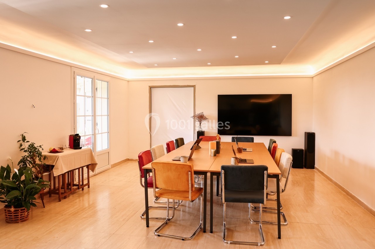 Salle de réunion lumineuse avec une grande table entourée de chaises colorées, écran mural et plantes décoratives.