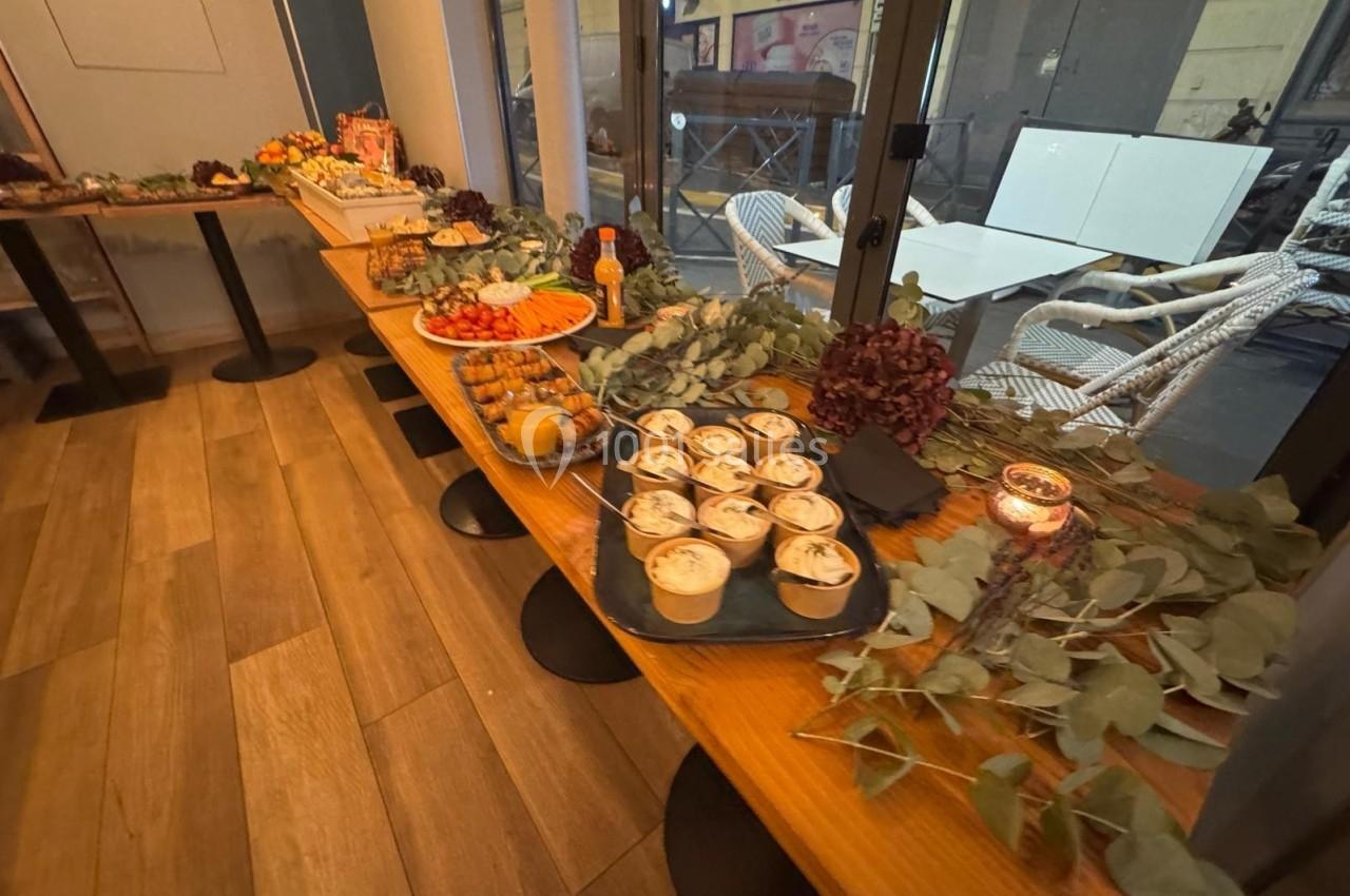 Table en bois garnie de desserts, fruits, boissons et décorations végétales dans un espace intérieur lumineux.