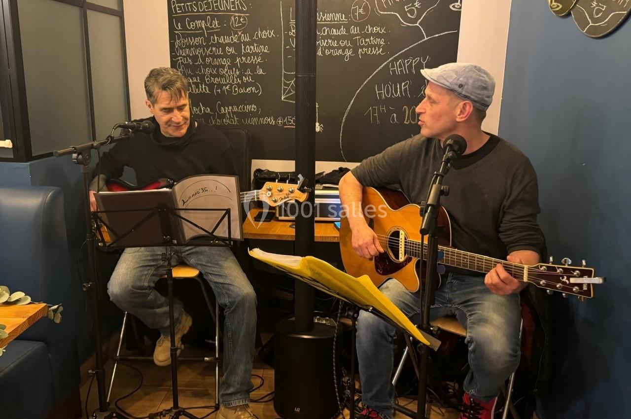 Deux musiciens jouent de la guitare dans un café, avec des partitions et un tableau noir en arrière-plan.