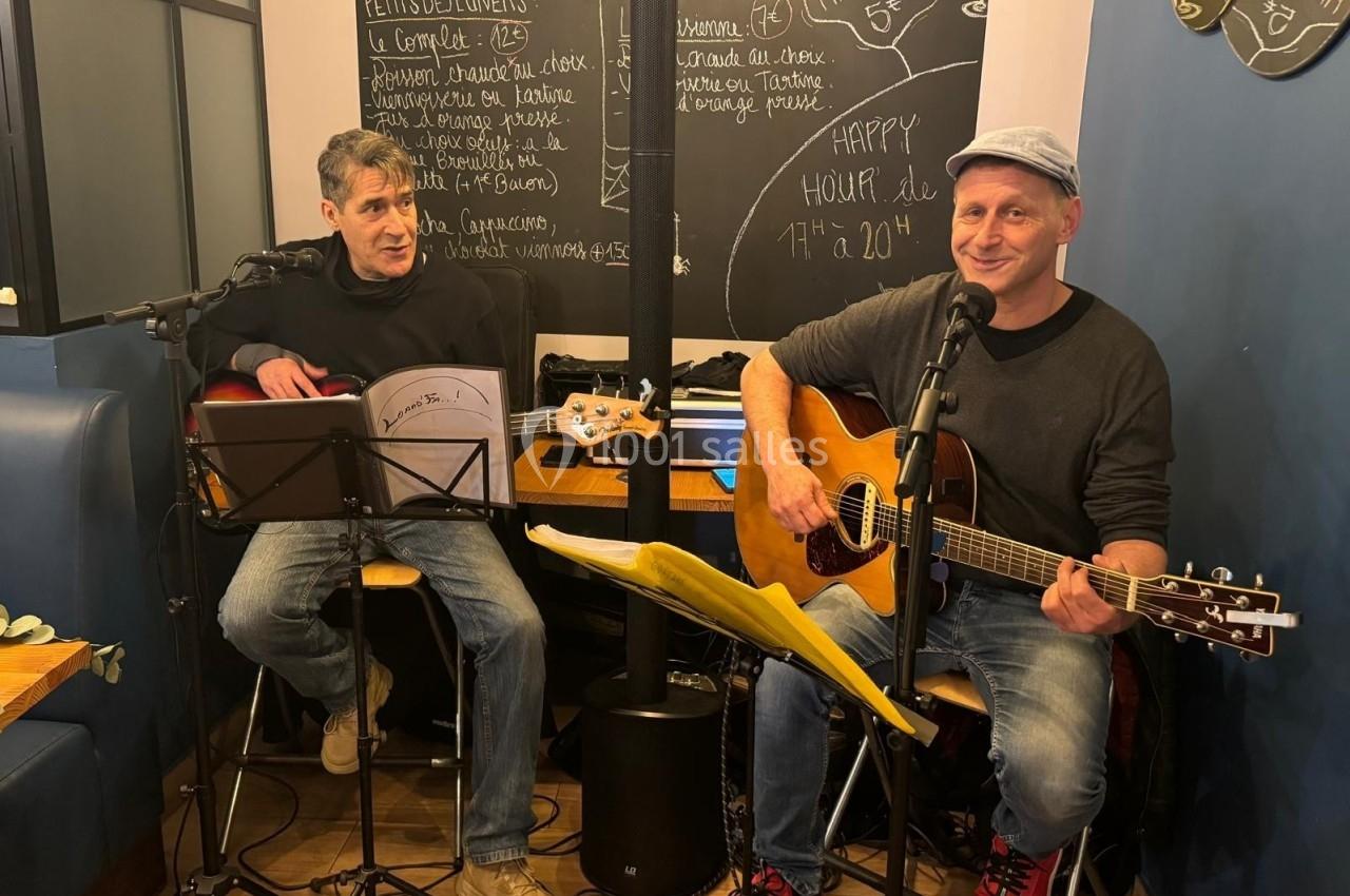 Deux musiciens jouent de la guitare et chantent dans un café, avec un tableau noir et des menus en arrière-plan.