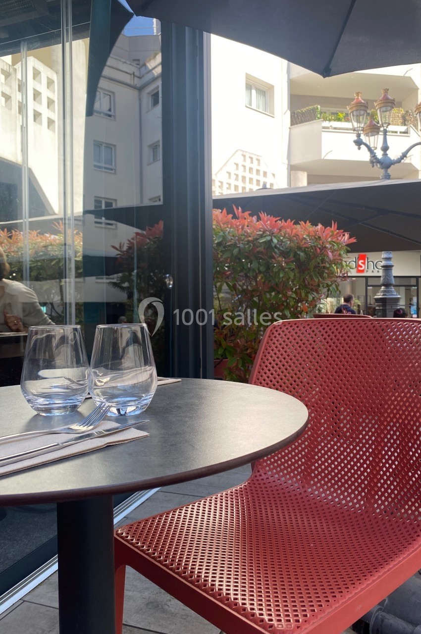 Table ronde avec deux verres et couverts sur une terrasse, chaise rouge et végétation en arrière-plan.