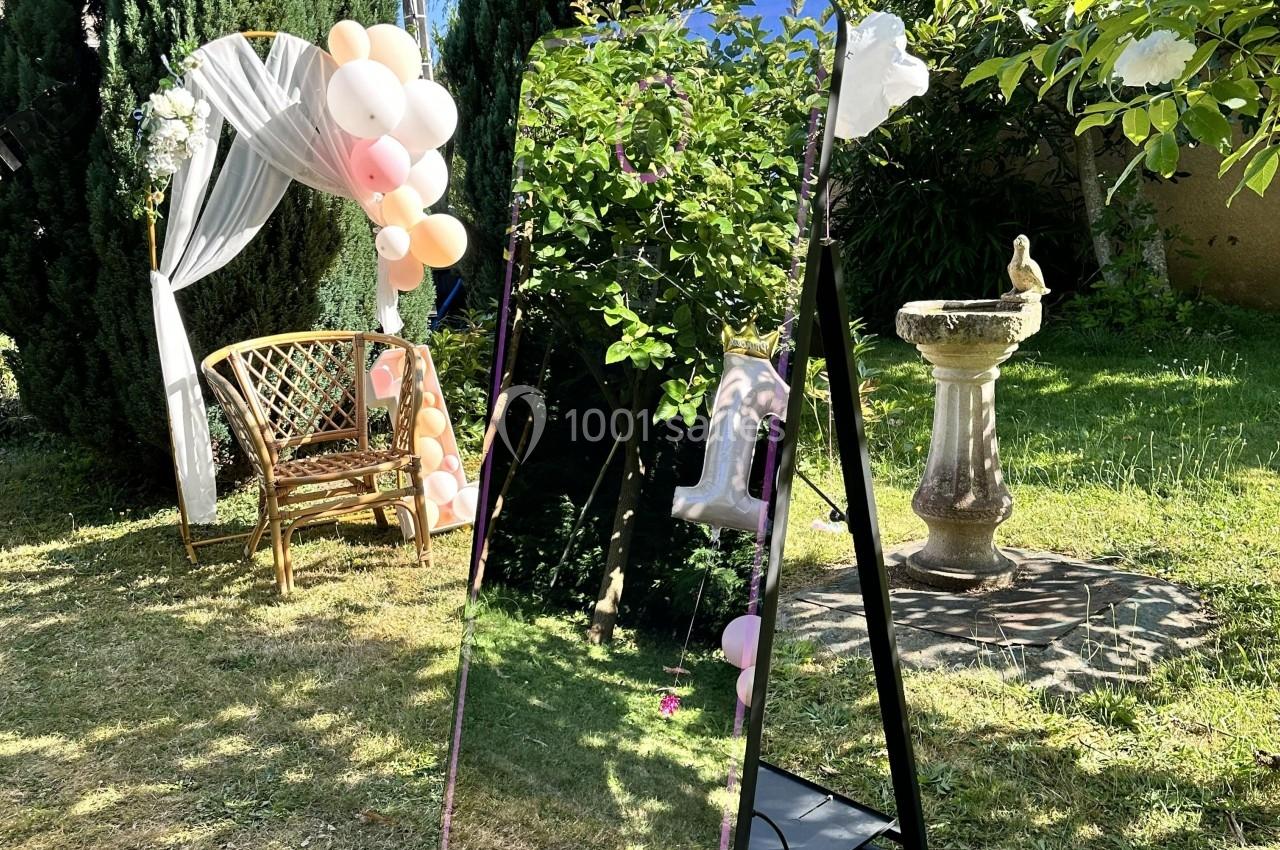 Miroir sur pied dans un jardin, entouré de verdure, avec une arche décorée de ballons et un banc en arrière-plan.