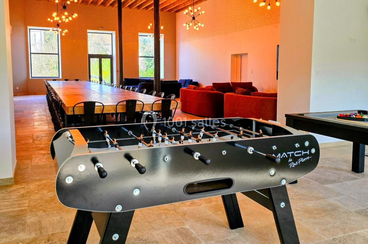 Baby-foot noir au premier plan dans une salle lumineuse avec une grande table, des canapés et un billard en arrière-plan.