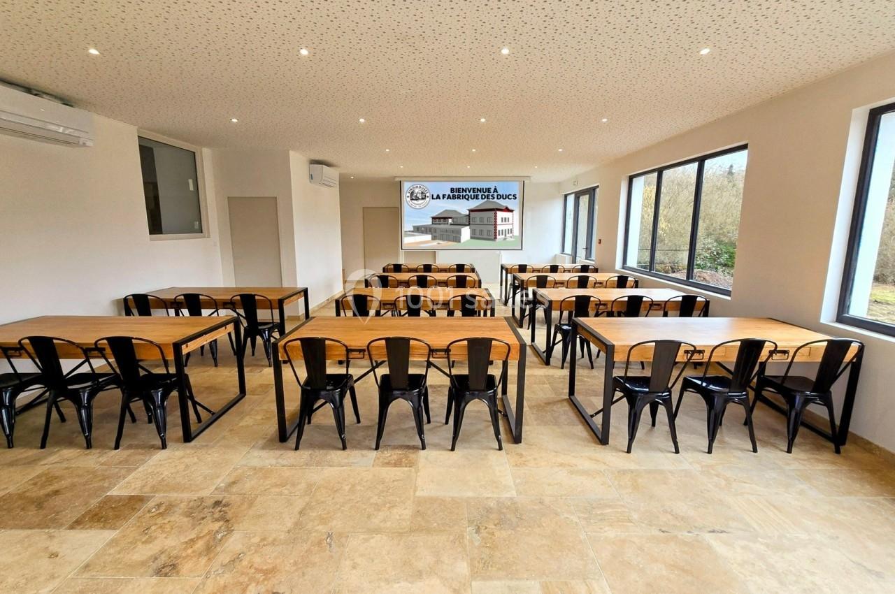 Salle lumineuse avec des tables en bois et des chaises noires, grandes fenêtres donnant sur un espace extérieur.