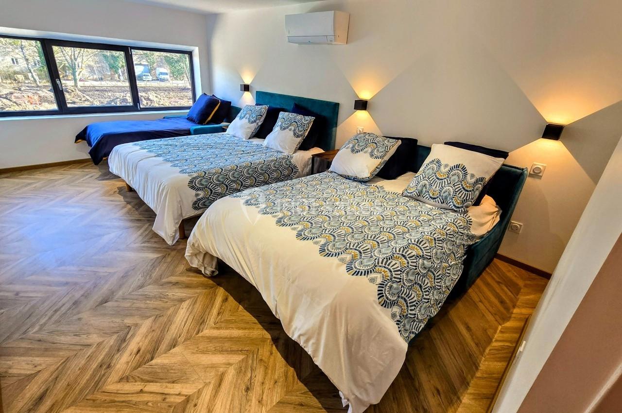 Trois lits simples alignés dans une chambre lumineuse avec parquet, éclairage mural et grande fenêtre donnant sur l…