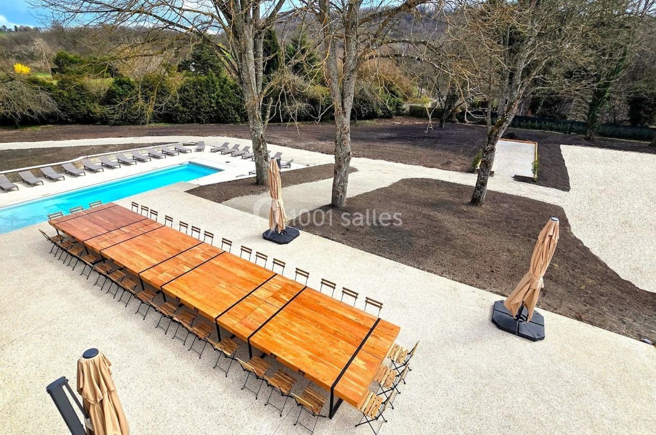 Terrasse extérieure avec longues tables en bois, parasols fermés, piscine et espaces verts en arrière-plan.