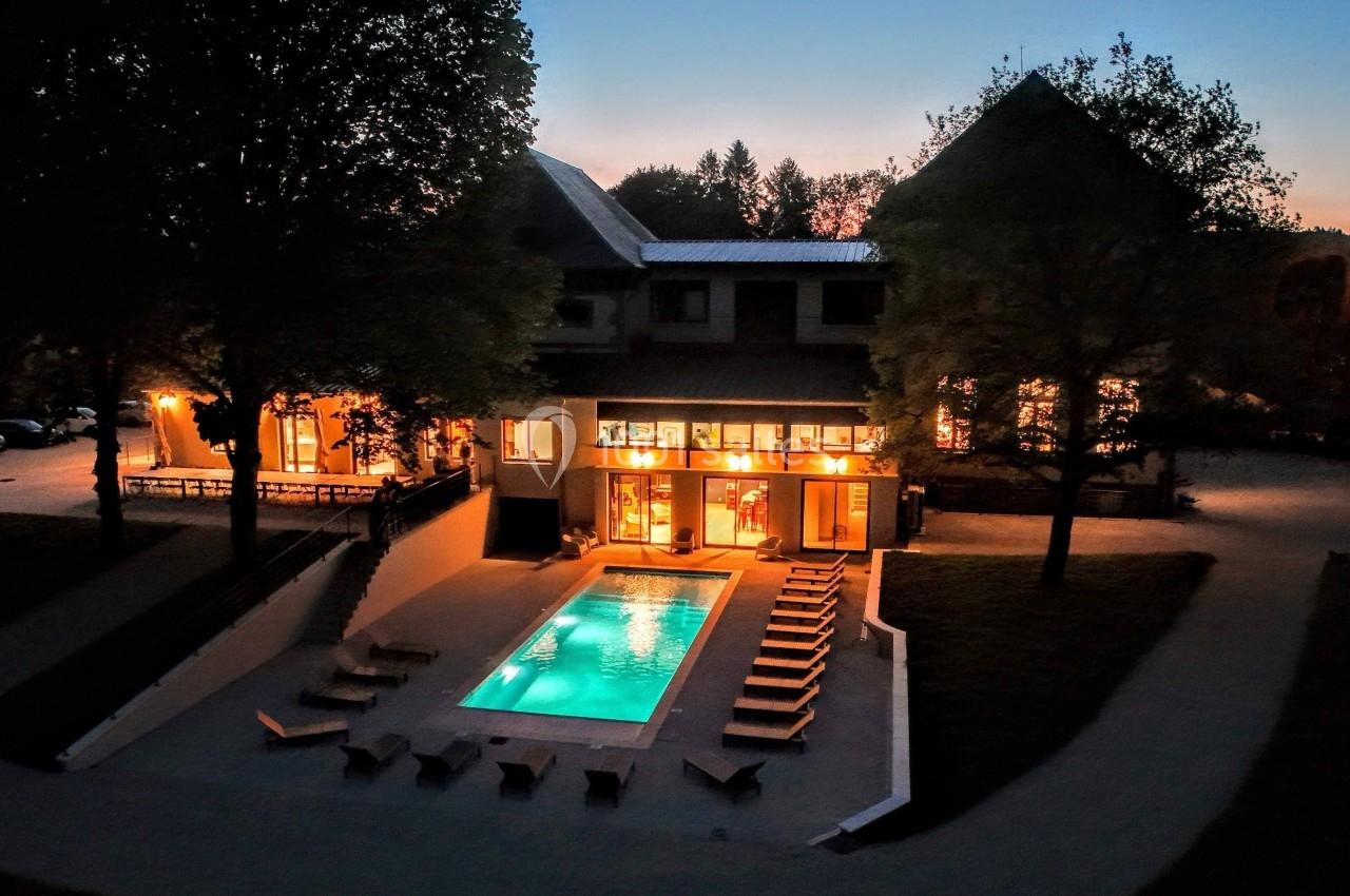 Vue nocturne d'une maison éclairée avec piscine extérieure entourée de chaises longues et d'arbres.