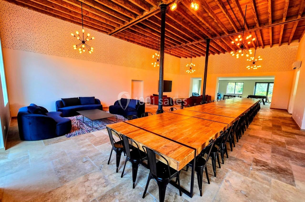 Salle spacieuse avec une grande table en bois, chaises noires, canapés bleus et plafond en bois avec luminaires suspendus.