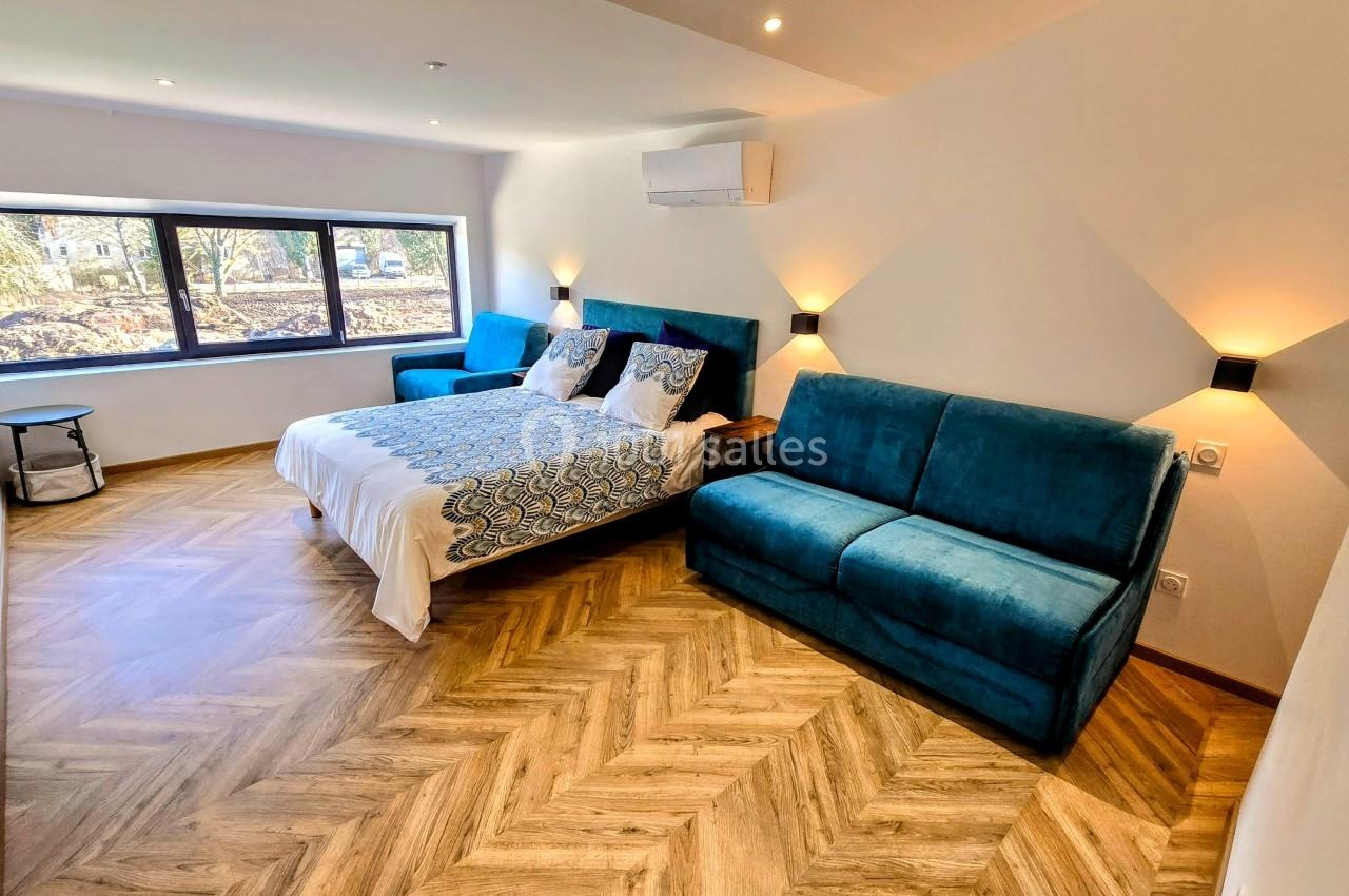 Chambre lumineuse avec lit double, canapé bleu, parquet en chevrons et grande fenêtre donnant sur un extérieur arboré.