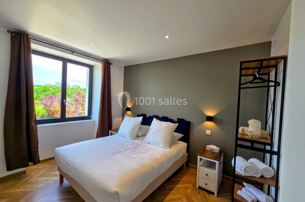 Chambre lumineuse avec lit double, draps blancs, murs neutres, fenêtre donnant sur un paysage verdoyant et parquet au sol.