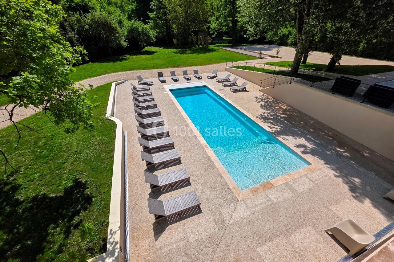 Piscine extérieure rectangulaire entourée de transats sur une terrasse, avec pelouse et arbres en arrière-plan.