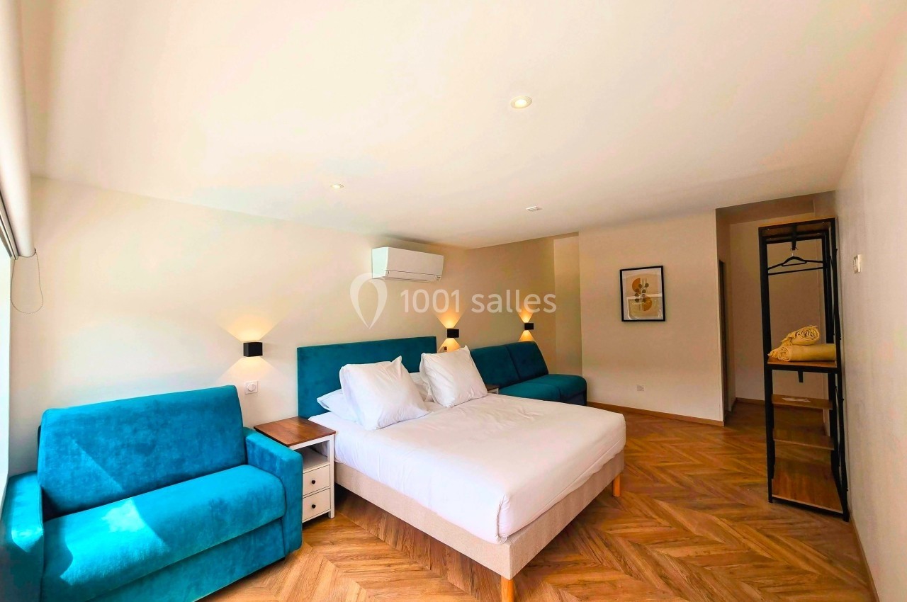 Chambre lumineuse avec lit double, deux canapés bleus, parquet en chevrons et éclairage mural.