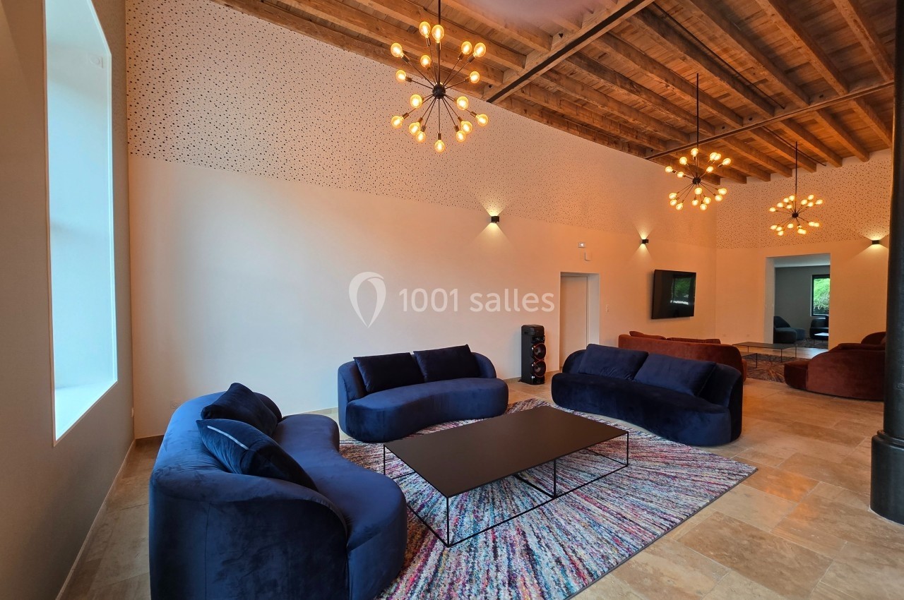 Salon spacieux avec canapés bleus, table basse noire, tapis coloré et plafond en bois avec luminaires suspendus.