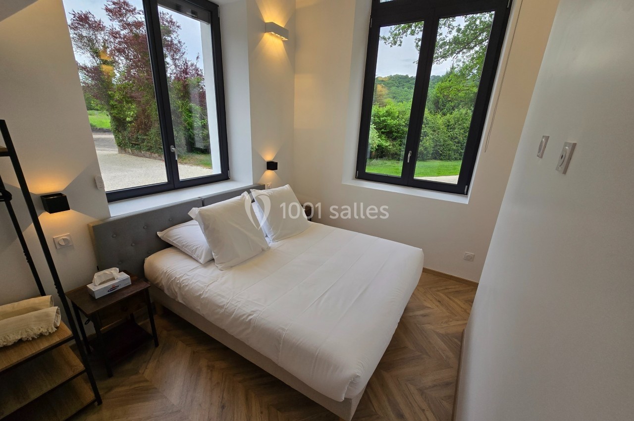 Chambre lumineuse avec lit double, parquet en bois, deux grandes fenêtres donnant sur un jardin verdoyant.