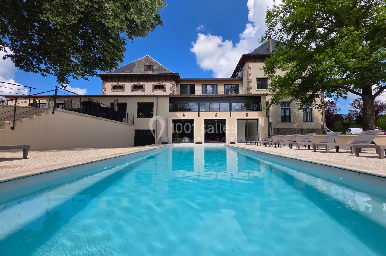 Piscine extérieure devant une grande maison en pierre avec terrasse, entourée d'arbres sous un ciel bleu.