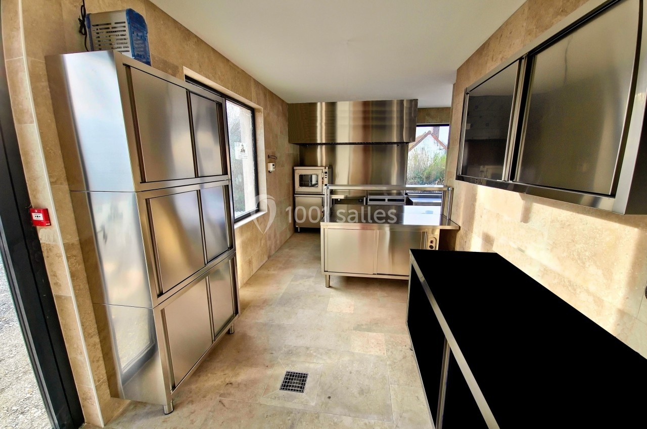 Cuisine professionnelle moderne avec équipements en acier inoxydable, murs en pierre beige et lumière naturelle.