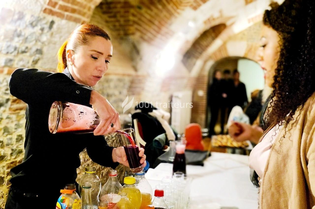 Une serveuse verse une boisson rouge dans un verre sur une table garnie de bouteilles, face à une invitée.