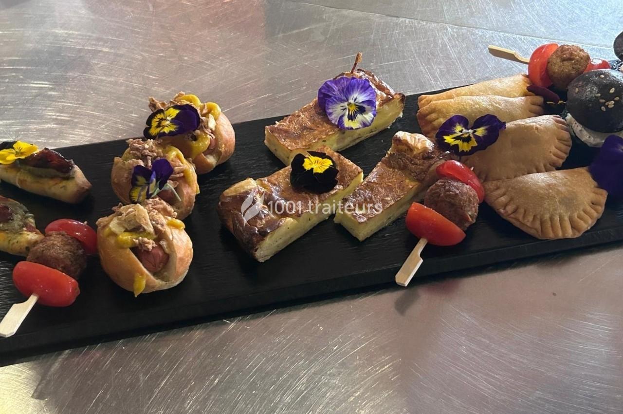 Assortiment de bouchées salées sur un plateau noir, incluant tartelettes, brochettes et feuilletés, décorés de fleurs…