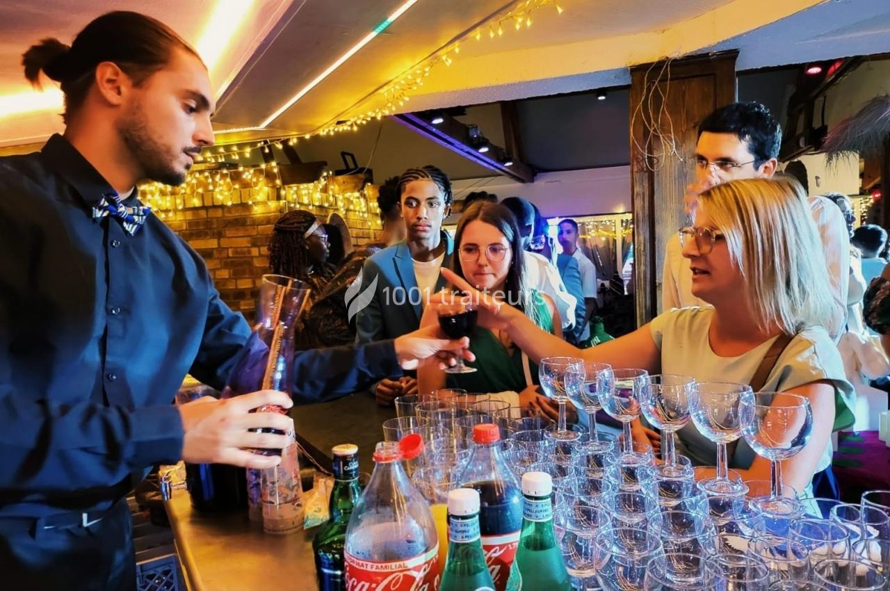 Un barman sert un verre de vin rouge à une femme lors d'un événement dans un bar éclairé par des guirlandes lumineuses.