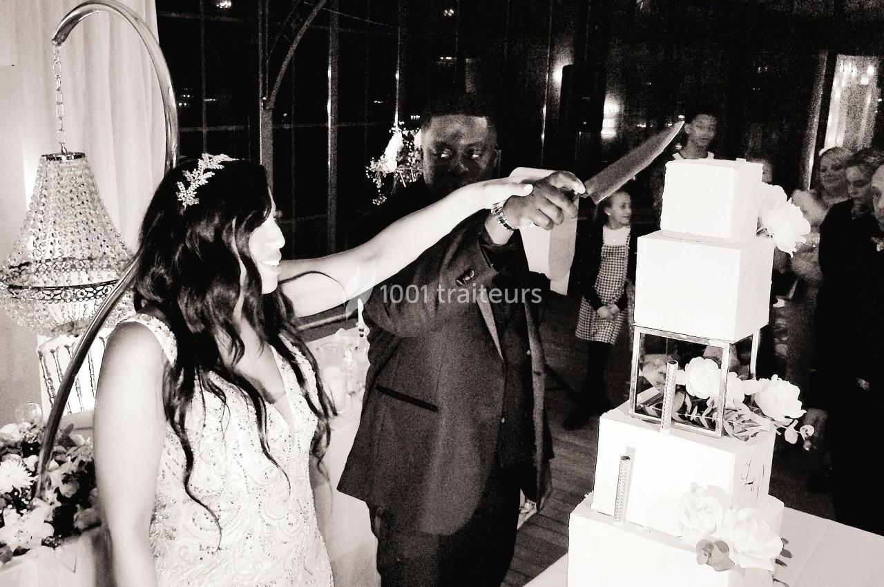 Un couple coupe un gâteau de mariage à plusieurs étages dans une salle décorée de fleurs et de lustres.