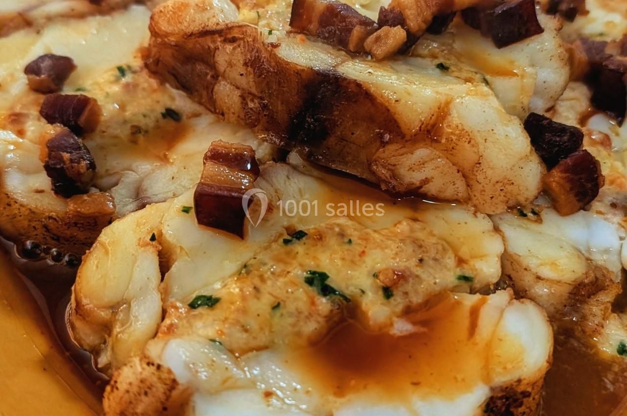 Tranches de lotte servies avec une sauce et des morceaux de lard sur un plat en terre cuite.