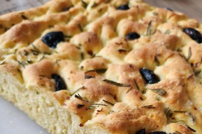 Focaccia dorée garnie de romarin et d'olives noires, posée sur une surface claire.