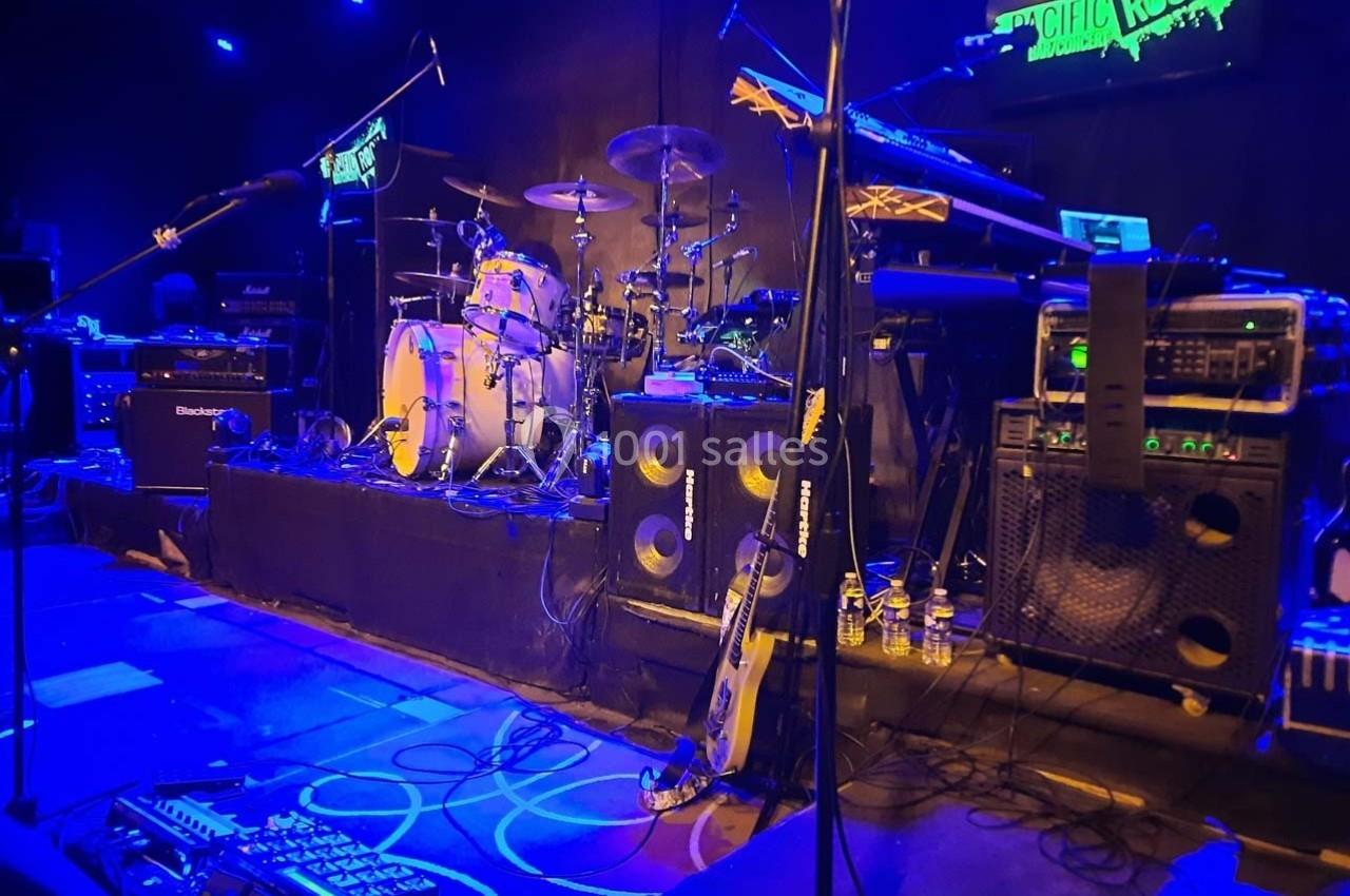 Scène de concert avec batterie, amplificateurs, guitares et éclairage bleu dans une salle de spectacle.