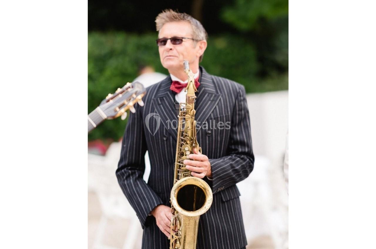 Un homme en costume rayé joue du saxophone en extérieur, avec une guitare visible en arrière-plan.
