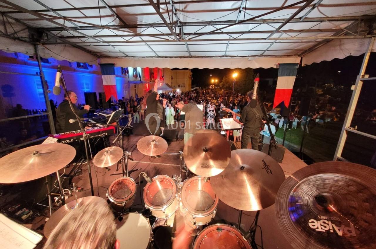 Vue depuis la scène d'un concert en plein air, montrant des musiciens et une foule sous des éclairages tricolores.