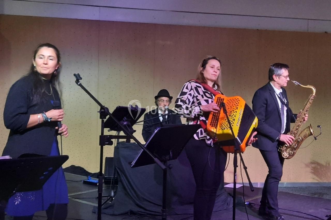 Un groupe de musiciens joue sur scène, avec une chanteuse, un accordéoniste, un saxophoniste et un pianiste.