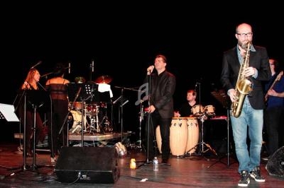 Un groupe de musiciens joue sur scène avec divers instruments, dont un saxophone, des percussions et une batterie.