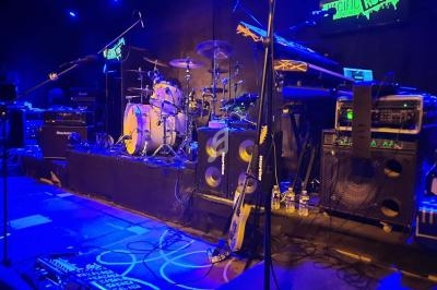 Scène de concert avec batterie, amplificateurs, guitares et éclairage bleu dans une salle de spectacle.