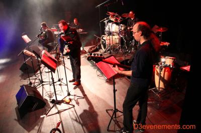 Un batteur joue sur une batterie argentée sur une scène sombre, avec un micro à proximité.