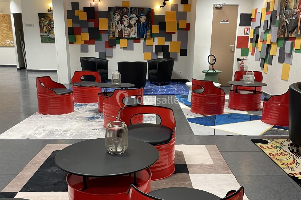 Espace lounge coloré avec tables et sièges en métal rouge, décoré de tableaux et de panneaux muraux géométriques.