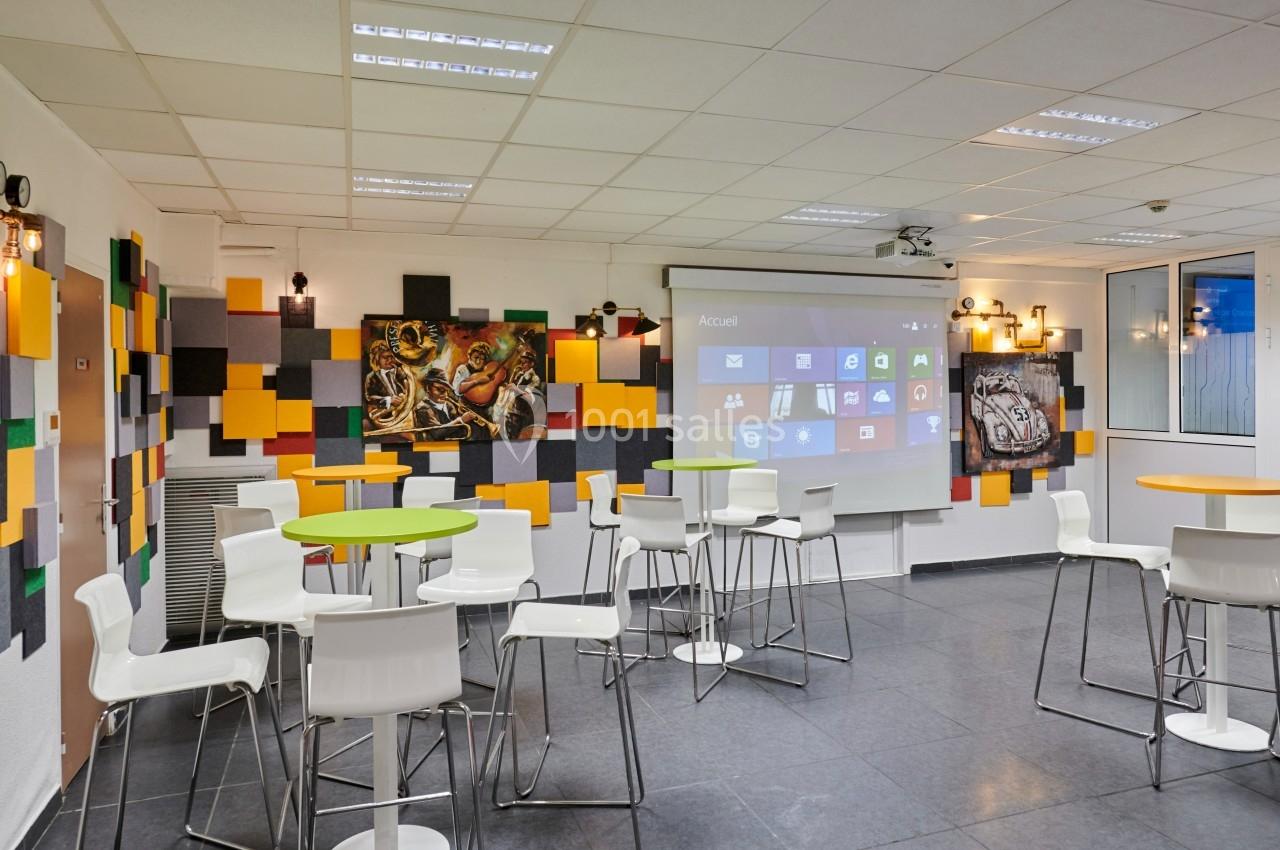 Salle moderne avec tables hautes, chaises blanches, écran de projection allumé et murs décorés de panneaux colorés.