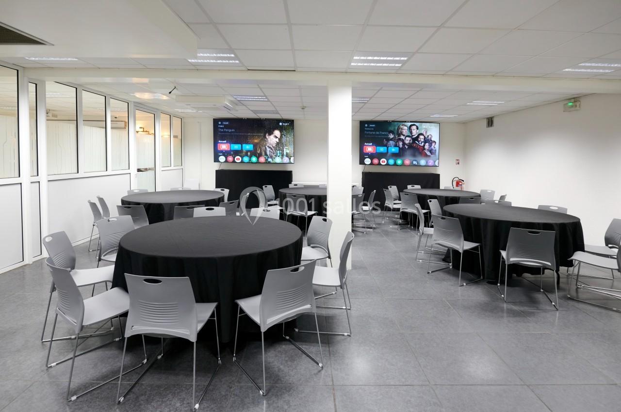 Salle de réunion avec tables rondes couvertes de nappes noires, chaises grises et écrans accrochés aux murs.