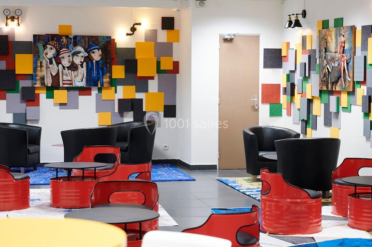 Espace lounge coloré avec fauteuils noirs, tables rouges, murs décorés de panneaux colorés et tableaux artistiques.