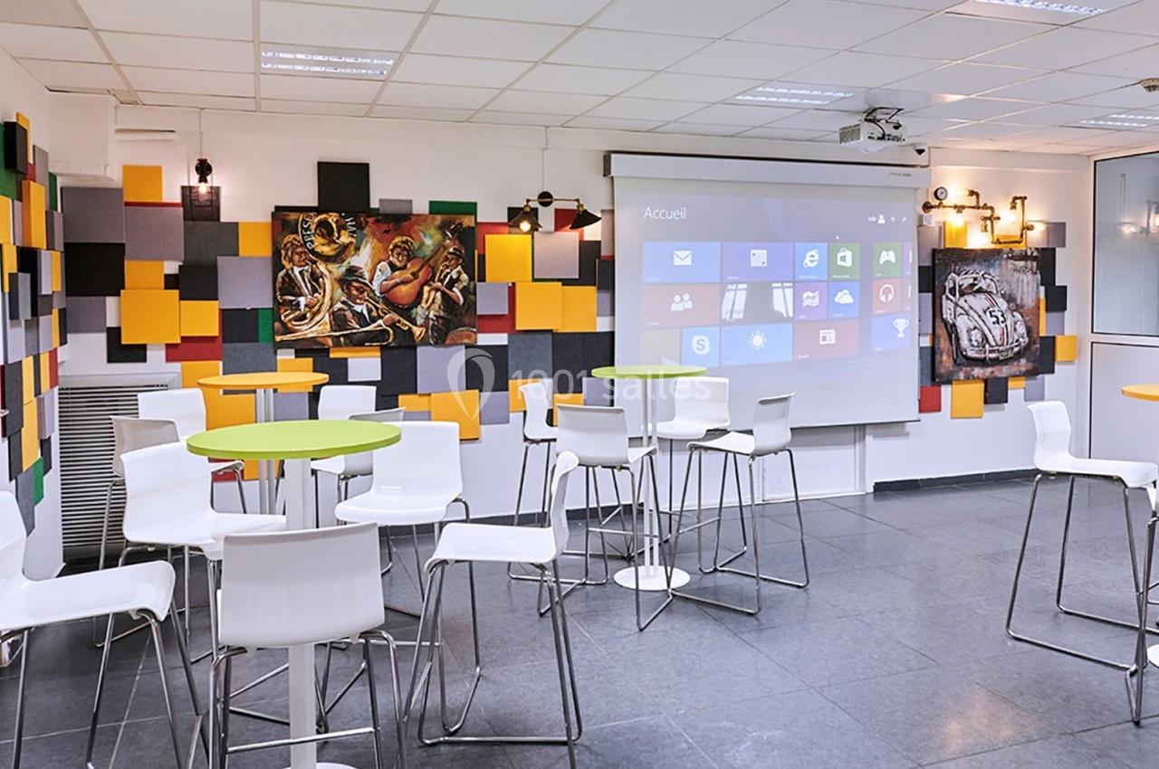 Salle moderne avec tables hautes, chaises blanches, décor coloré au mur et écran de projection allumé.