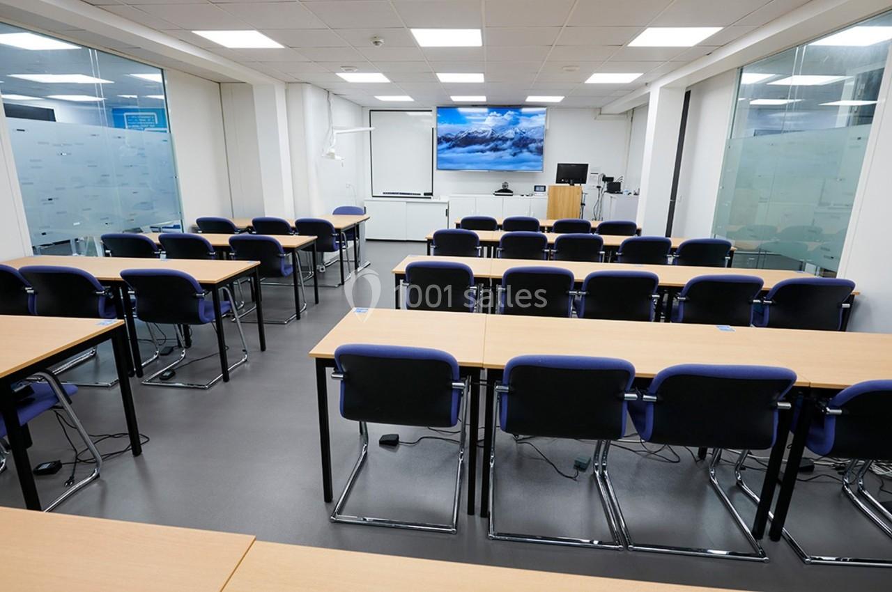 Salle de formation vide avec des rangées de tables et chaises, écran de projection allumé au fond de la pièce.