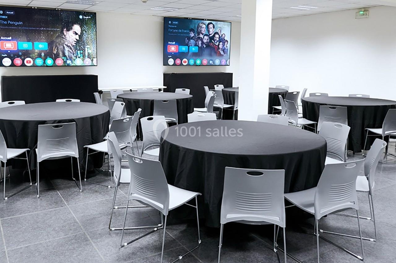 Salle de réunion avec plusieurs tables rondes noires, entourées de chaises grises, et deux écrans accrochés au mur.