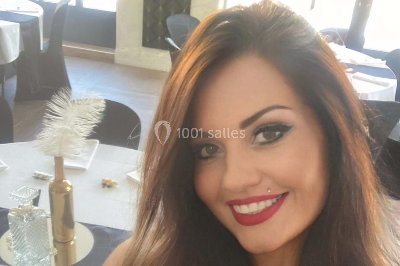 Une femme souriante avec des cheveux bruns, posant dans une salle de réception élégante avec des tables décorées.