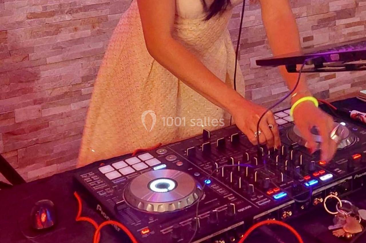 Une femme en robe claire mixe de la musique sur une table de mixage avec des câbles et des lumières colorées.