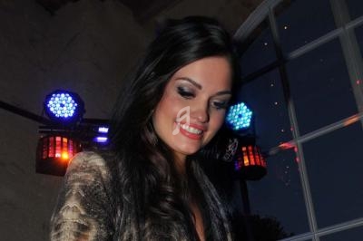 Femme souriante avec des cheveux bruns longs, éclairée par des lumières colorées dans un intérieur avec une grande fenêtre.